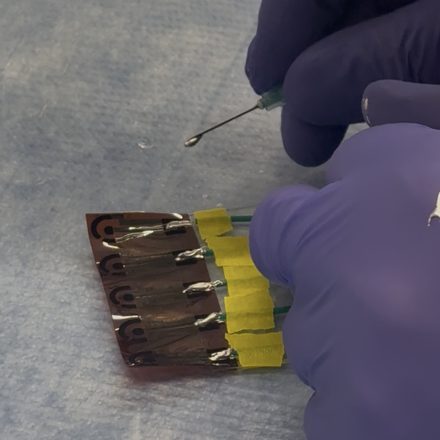 A gloved hand holding a syringe over a flexible electrode array, its five leads taped down to the bench with yellow tape